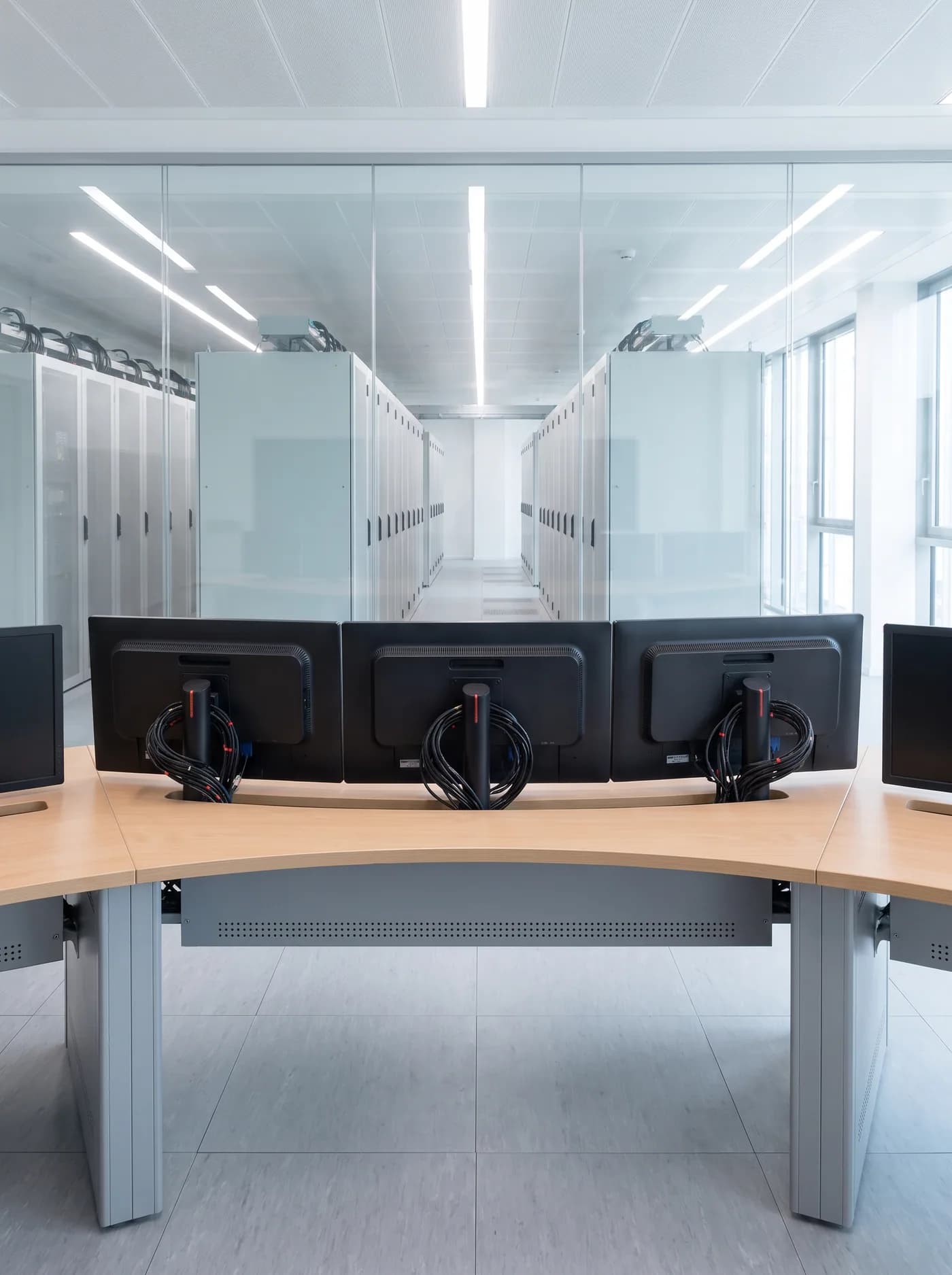 A network operations room seen from behind an empty desk: the backs of a bank of monitors and their cable looms, and beyond a glass wall a long aisle of cabinets in the hall below.