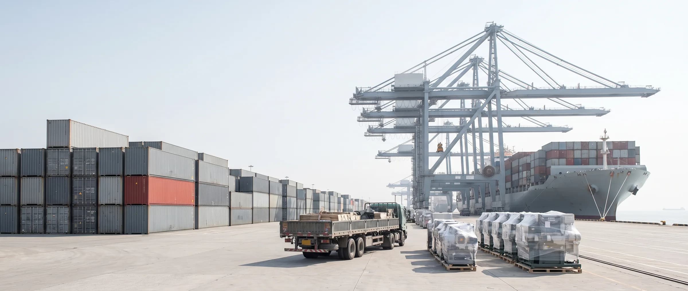 A Chinese container terminal at the moment of export: stacked containers running to the horizon, gantry cranes over a moored ship, and palletised machinery waiting on the apron.